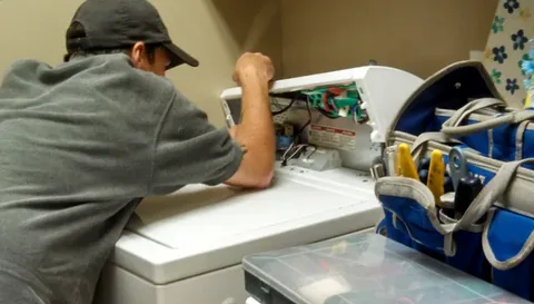 A technician wearing a black cap and gray shirt repairs a white washing machine with its top panel open beside a blue tool bag, addressing Common Appliance Problems such as drainage issues, spinning failures, and washer performance concerns.