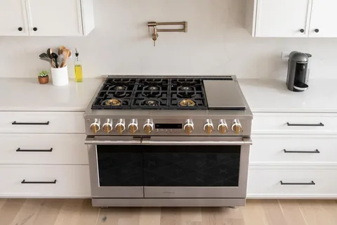 A stainless steel gas range with a built-in griddle is positioned between white cabinets and drawers in a modern kitchen, featuring a gold wall-mounted pot filler faucet, showcasing a well-maintained cooking area often supported by professional appliance repair services.